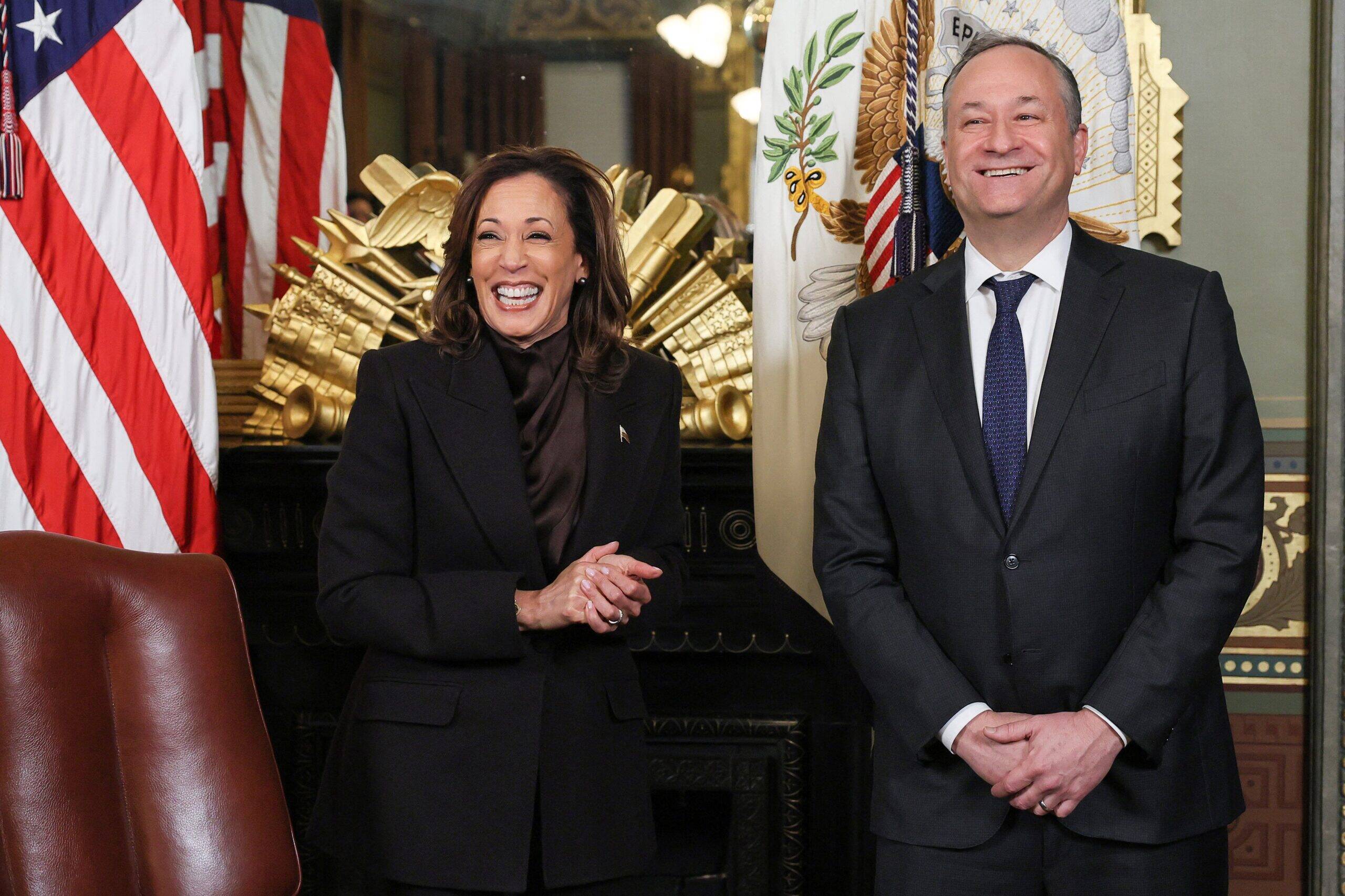 Vice President Kamala Harris stands smiling beside her husband Doug Emhoff in front of U.S. flags during an official event.